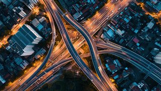 Luftaufnahme eines komplexen Autobahnkreuzes in einer Stadt bei Nacht. Mehrere mehrspurige Straßen verlaufen auf unterschiedlichen Ebenen und sind von beleuchteten Straßenlaternen gesäumt. Um das Autobahnkreuz herum befinden sich dicht bebaute Stadtviertel mit Wohn- und Geschäftsgebäuden. Die Lichter der fahrenden Autos erzeugen helle Lichtspuren, die die Dynamik des Verkehrs verdeutlichen.