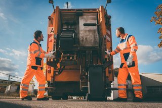 Zwei Müllwerker in orangefarbener Warnkleidung entleeren bei Tageslicht eine schwarze Mülltonne am Heck eines Müllwagens auf einer städtischen Straße.