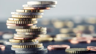 Schräg gestapelter Turm aus unterschiedlich großen Euro-Münzen in Silber, Gold und Kupfer. Im unscharfen Hintergrund liegen weitere Münzen verstreut auf dunkler Oberfläche. Symbolbild für Finanzen, Sparen oder wirtschaftliches Gleichgewicht.