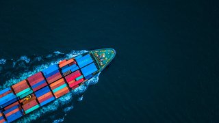 Luftaufnahme eines Containerschiffs, das mit bunten Frachtcontainern beladen über das offene Meer fährt und eine weiße Heckwelle hinter sich zieht.