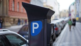 Das Bild zeigt einen Parkscheinautomaten am Straßenrand in einer innerstädtischen Umgebung. Auf der Vorderseite des Automaten ist ein blaues Parkschild mit weißem Buchstaben ‚P‘ zu sehen. Entlang der Straße stehen mehrere geparkte Autos, im Hintergrund sind mehrgeschossige Gebäude und unscharf erkennbare Passantinnen und Passanten auf dem Gehweg zu sehen.