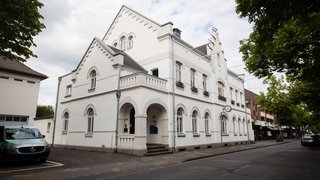 Das Rathaus Meerbusch: Ein helles, historisches Gebäudes an einer Straße mit Bäumen. Das zweigeschossige Haus ist weiß verputzt, hat Rundbogenfenster, dekorative Ziegelverzierungen und eine kleine Arkadenvorhalle. An der Ecke hängt eine runde Uhr, und im Vordergrund steht ein geparktes Fahrzeug. Neben dem Gebäude sind weitere Geschäfte und Häuser zu sehen.