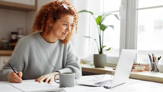 Junge Frau mit lockigen roten Haaren sitzt an einem Schreibtisch, arbeitet am Laptop und macht sich handschriftliche Notizen. Sie trägt einen grauen Strickpullover, vor ihr steht eine Tasse, im Hintergrund sind ein Fenster, eine Topfpflanze und Schreibutensilien zu sehen.