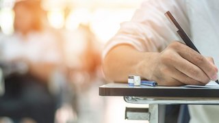 Person sitzt an einem Tisch und schreibt mit einem Stift auf Papier, im Hintergrund sind weitere unscharfe Personen in einem Klassenraum oder Prüfungsraum zu sehen.