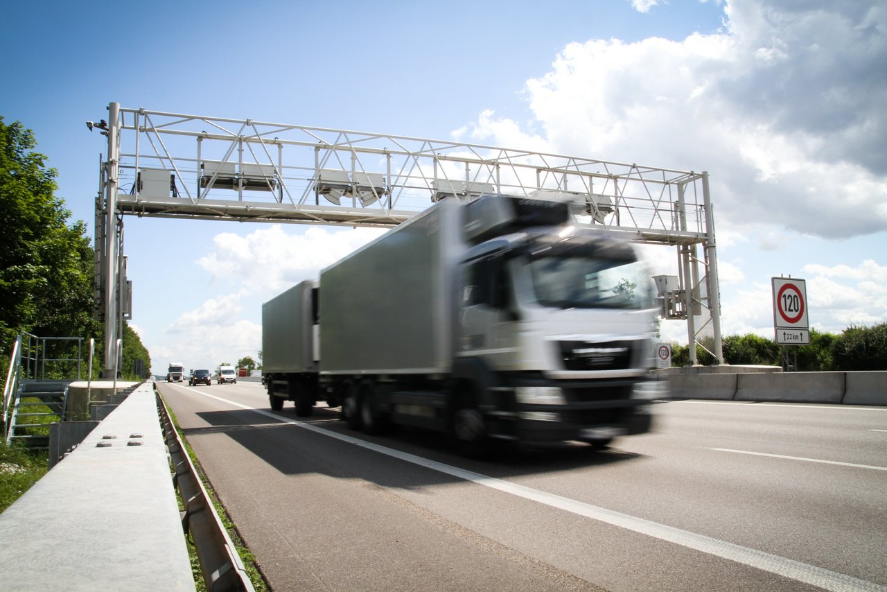 Ein LKW fährt mit hoher Geschwindigkeit auf einer mehrspurigen Autobahn unter einer Maut- oder Kontrollbrücke hindurch. Im Hintergrund sind weitere Fahrzeuge zu sehen. Rechts steht ein Verkehrszeichen mit einer Geschwindigkeitsbegrenzung von 120 km/h. Der Himmel ist teilweise bewölkt.