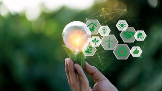 Nahaufnahme einer Hand, die eine leuchtende Glühbirne mit grünen Blättern darunter hält, vor einem verschwommenen, natürlichen Hintergrund. Rechts im Bild schweben sechseckige Symbole mit grünen Icons zu nachhaltiger Energie, Recycling, Solarenergie, Windkraft, Umweltschutz und Zusammenarbeit in einem digitalen Netzwerk.