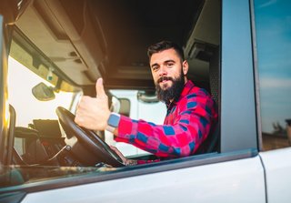 Ein Lkw-Fahrer mit Bart und kariertem Hemd sitzt in der Fahrerkabine eines Lastwagens, schaut aus dem offenen Fenster und zeigt mit einem Lächeln den Daumen nach oben. Das Sonnenlicht fällt von der Seite in die Kabine.