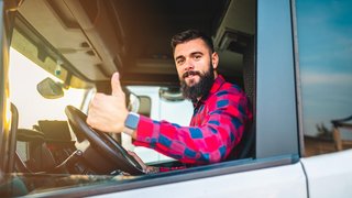 Ein Lkw-Fahrer mit Bart und kariertem Hemd sitzt in der Fahrerkabine eines Lastwagens, schaut aus dem offenen Fenster und zeigt mit einem Lächeln den Daumen nach oben. Das Sonnenlicht fällt von der Seite in die Kabine.