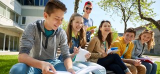 Eine Gruppe von sieben Jugendlichen sitzt bei sonnigem Wetter auf einer Holzbank im Freien vor einem modernen Schul- oder Universitätsgebäude. Die Jugendlichen lachen, unterhalten sich und halten Hefte oder Schreibmaterialien in der Hand. Die Atmosphäre wirkt freundlich, entspannt und gemeinschaftlich.