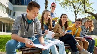 Eine Gruppe von sieben Jugendlichen sitzt bei sonnigem Wetter auf einer Holzbank im Freien vor einem modernen Schul- oder Universitätsgebäude. Die Jugendlichen lachen, unterhalten sich und halten Hefte oder Schreibmaterialien in der Hand. Die Atmosphäre wirkt freundlich, entspannt und gemeinschaftlich.