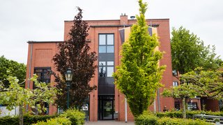 Das Bild zeigt das Rathaus von Korschenbroich von vorne. Zu sehen ist ein mehrgeschossiges Verwaltungsgebäude aus rotem Backstein mit einem zentralen, hohen Eingangsbereich und großen, vertikal angeordneten Fenstern. Vor dem Gebäude führt ein gepflasterter Weg zum Eingang, eingerahmt von Büschen und mehreren Bäumen mit unterschiedlich gefärbtem Laub. Das Rathaus ist von Grünflächen umgeben, der Himmel ist hell und bewölkt.