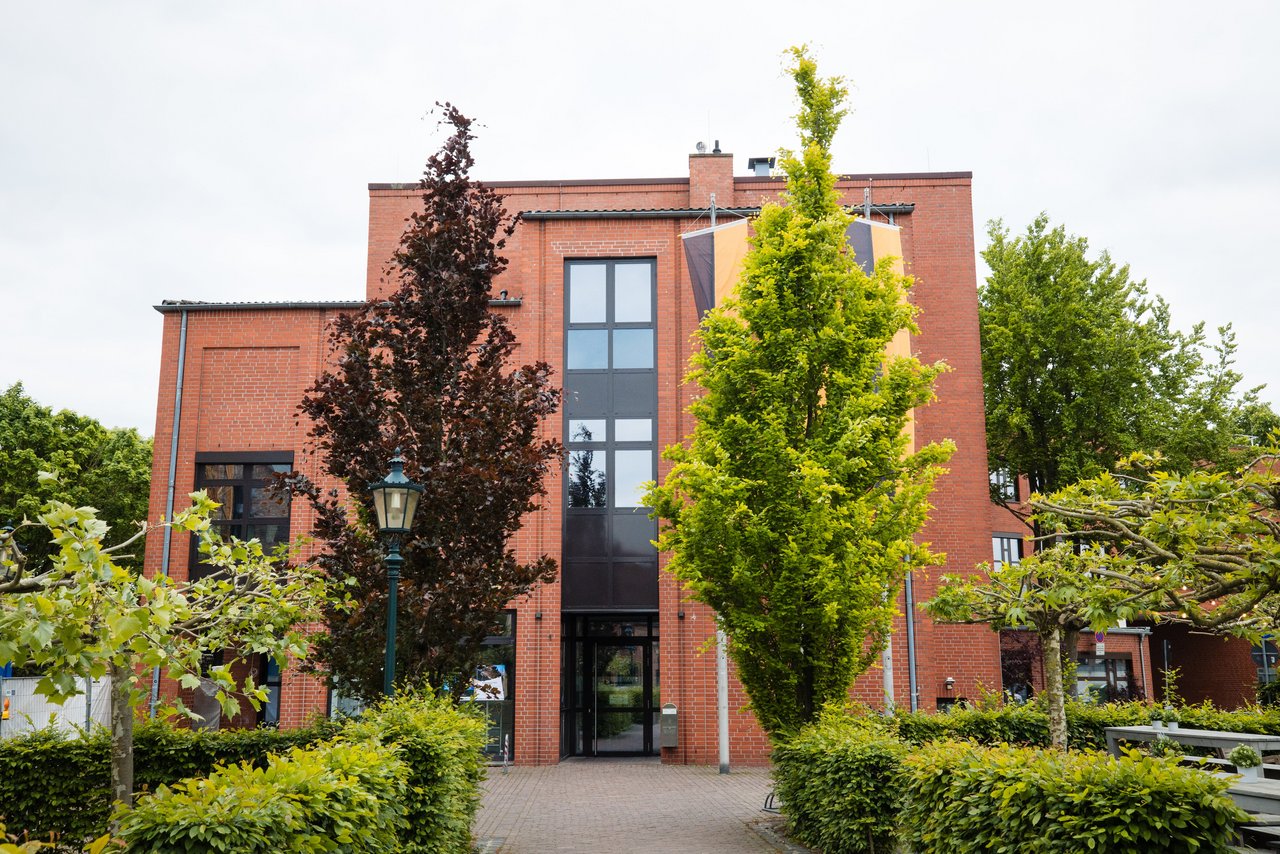 Das Bild zeigt das Rathaus von Korschenbroich von vorne. Zu sehen ist ein mehrgeschossiges Verwaltungsgebäude aus rotem Backstein mit einem zentralen, hohen Eingangsbereich und großen, vertikal angeordneten Fenstern. Vor dem Gebäude führt ein gepflasterter Weg zum Eingang, eingerahmt von Büschen und mehreren Bäumen mit unterschiedlich gefärbtem Laub. Das Rathaus ist von Grünflächen umgeben, der Himmel ist hell und bewölkt.