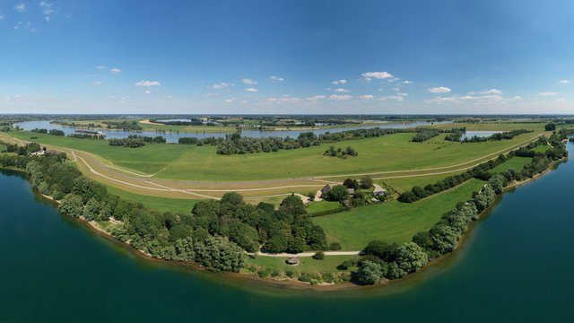 Luftaufnahme einer weitläufigen Seenlandschaft, der Xantener Nordsee, mit grünen Wiesen, Feldern und kleinen Wäldern. Im Vordergrund liegt eine Landzunge, die von Wasser umgeben ist, mit vereinzelten Gebäuden und einem Weg entlang des Ufers. Im Hintergrund erstrecken sich weitere Seen, Wiesen und kleine Ortschaften unter einem blauen Himmel mit wenigen Wolken.